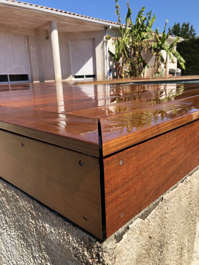 Coin d'un pont de piscine mobile en bois exotique (IPE) fraîchement huilé, reflétant la maison et les bananiers sous le soleil.