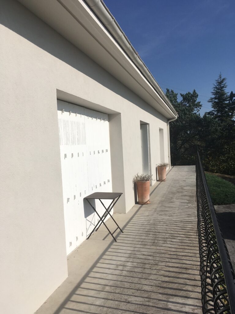 Façade moderne blanche avec porte de garage à lattes. Patio en béton, ombre marquée d'une balustrade en fer forgé. Petite table noire.